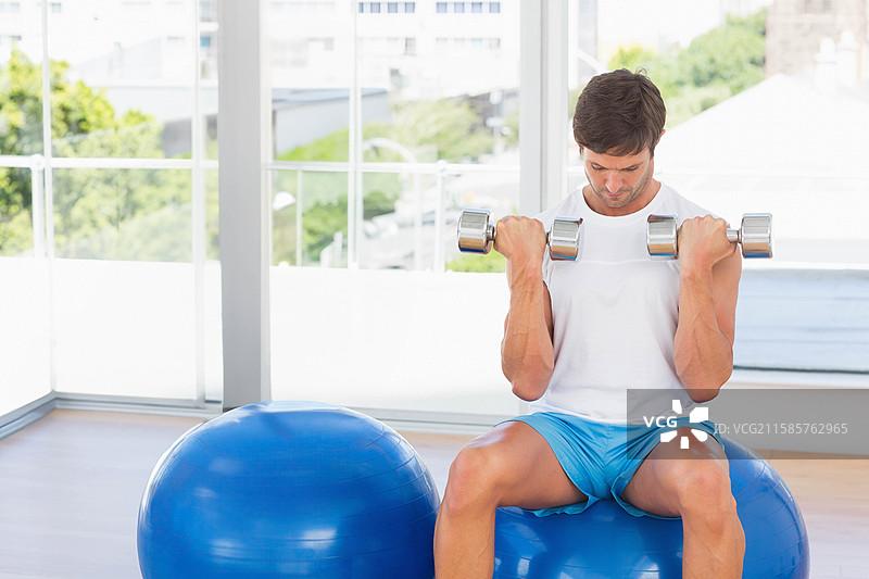 穿着运动服的男子坐在稳定球上，双手持哑铃于肩部高度，健身房内有空白空间。图片素材