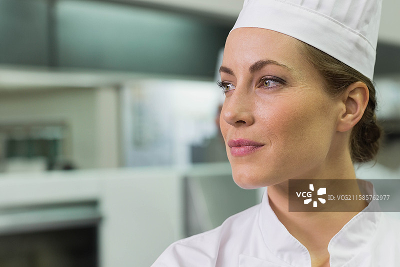 中年女性厨师站在厨房的不锈钢台面旁，戴着厨师帽和外套，留有空白空间图片素材