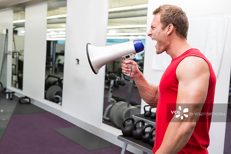 在健身房的举重区，一名男子手持扩音器，靠近壶铃和哑铃大声喊叫，旁边留有空白空间。图片素材