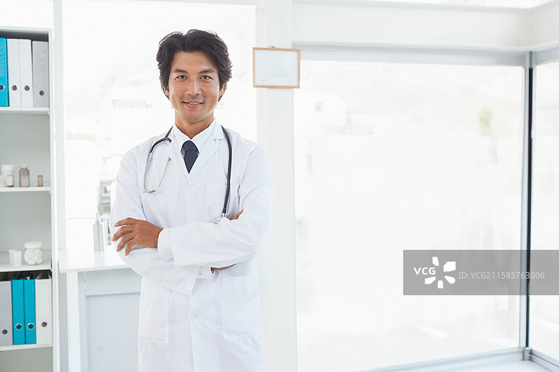穿着白大褂和听诊器的日本男性医生，双臂交叉站在诊所的货架旁。图片素材