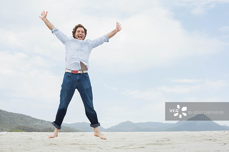赤脚男子穿着轻便衬衫和卷起的牛仔裤，在沙滩上跳跃，云层笼罩。图片素材