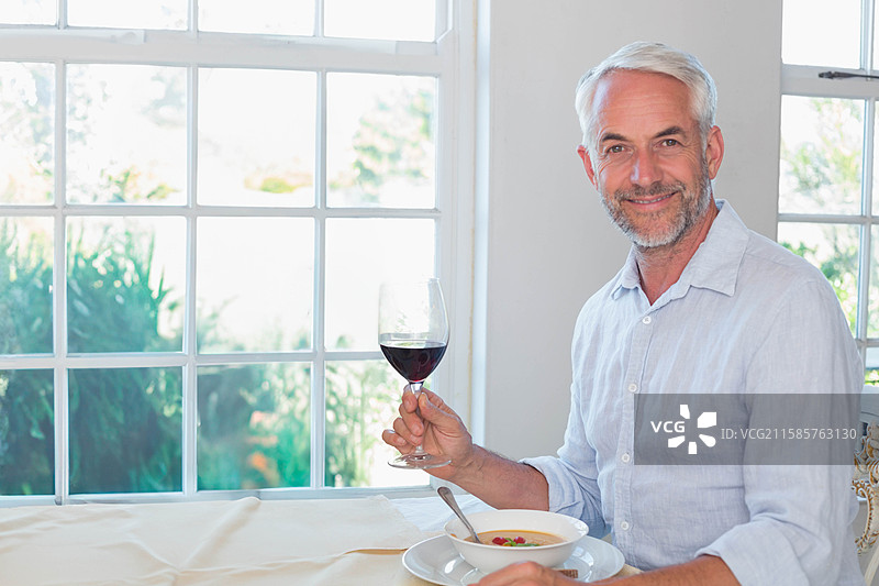 成熟男性坐在窗边的餐桌旁，手里拿着酒杯和汤碗，留有空白空间图片素材