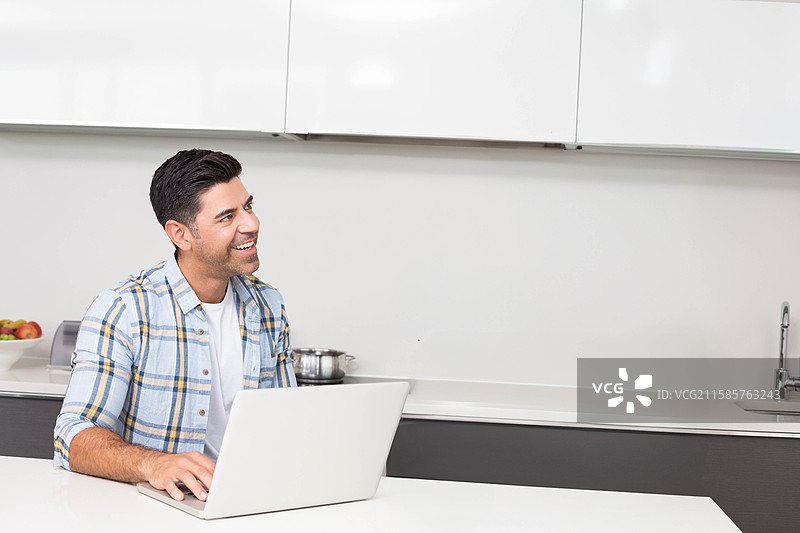 中年男子在厨房岛台上使用笔记本电脑，旁边有一碗苹果和一个炊具锅。图片素材