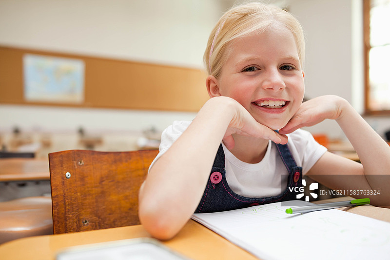 女童坐在教室的木制书桌前，微笑着手握笔记本和钢笔，留有空白空间。图片素材