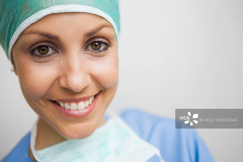 女性医疗工作者微笑着，戴着绿色手术帽和口罩，穿着蓝色医护服，在诊所里。图片素材