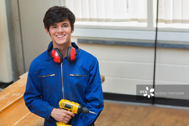 穿着工作服、戴着红色耳罩的男性技术员在车间的工作台上微笑着使用黄色电钻。图片素材