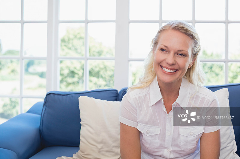 阳光明媚的客厅里，女人坐在蓝色沙发上，旁边有白色靠垫，窗户为多窗格设计，留有空白空间。图片素材