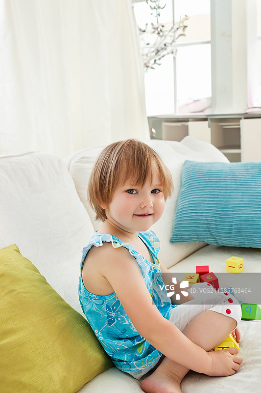 坐在客厅白色沙发上的小女孩盘腿而坐，周围是五颜六色的字母积木。图片素材
