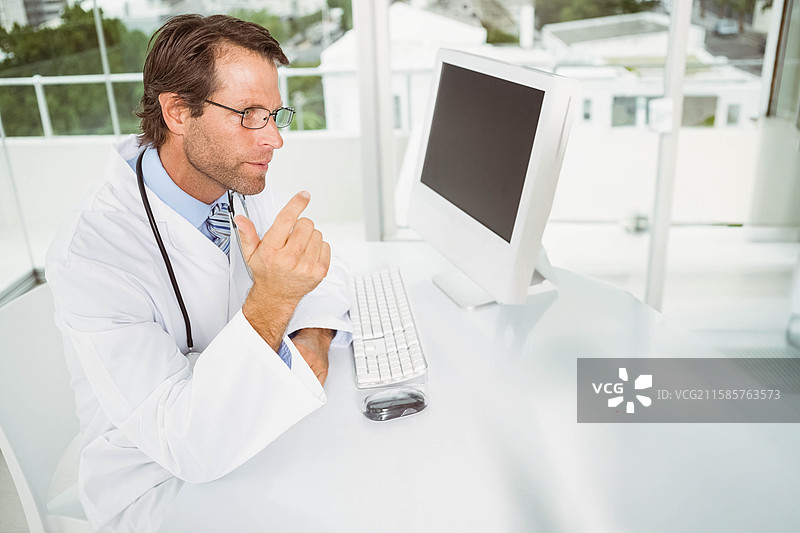 穿着白大褂和听诊器的医生坐在桌子前，指着显示器，留有空白空间图片素材