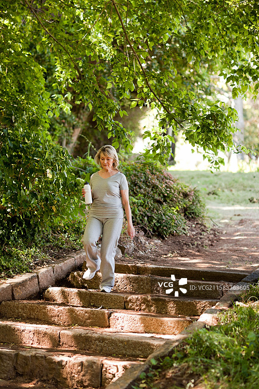穿着灰色运动服的老年女性在公园的石阶上走下，手里拿着白色水瓶。图片素材