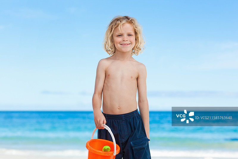 金发男孩站在海滩上，手里拿着一个橙色塑料桶和一把绿色玩具铲子，海浪拍打着岸边。图片素材