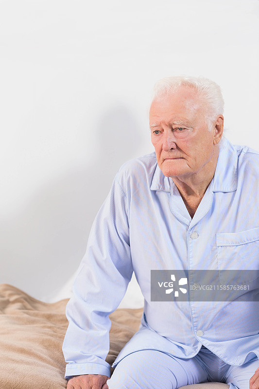 穿着睡衣的老年人靠在床边，旁边是米色的毯子和床垫，留有空白空间。图片素材