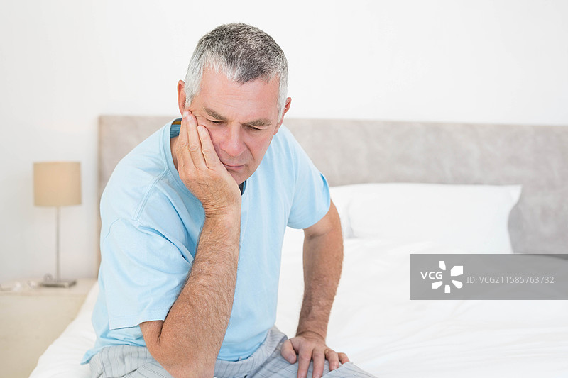 穿着睡衣的老年男子坐在床上，脸颊靠在床头柜的台灯旁，留有空白空间。图片素材