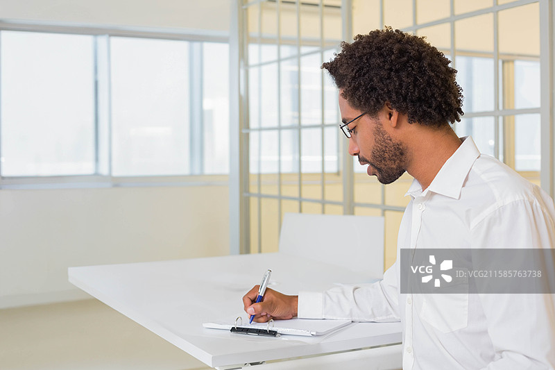 办公室专业人士靠在白色桌子上，正在玻璃隔断旁的窗户附近的夹板上写字。图片素材