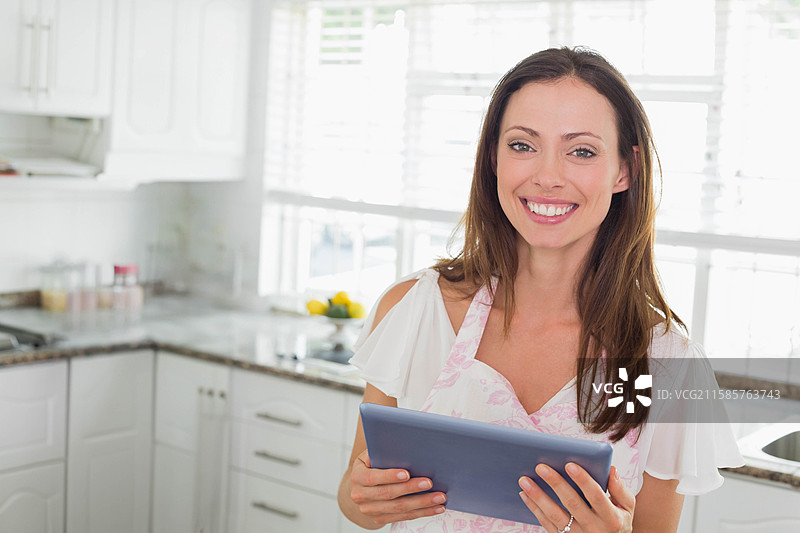 穿着花卉围裙的女人在厨房的台面上拿着平板电脑查看食谱，旁边有香料罐。图片素材