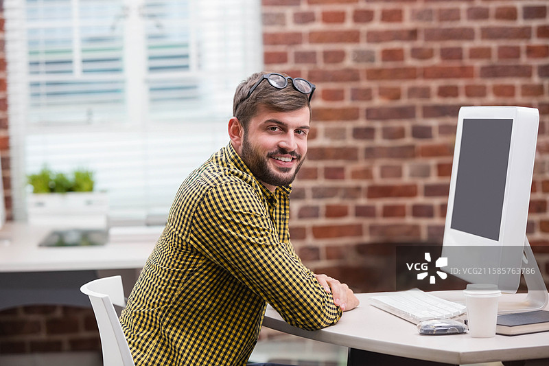 戴眼镜的男子微笑着靠在办公室的桌子上，桌上有电脑和咖啡杯，背景是砖墙。图片素材
