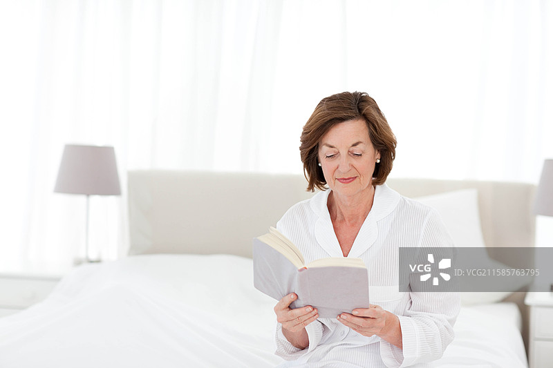 老年女性坐在卧室窗边的床上阅读平装书，旁边有台灯，留有空白空间。图片素材