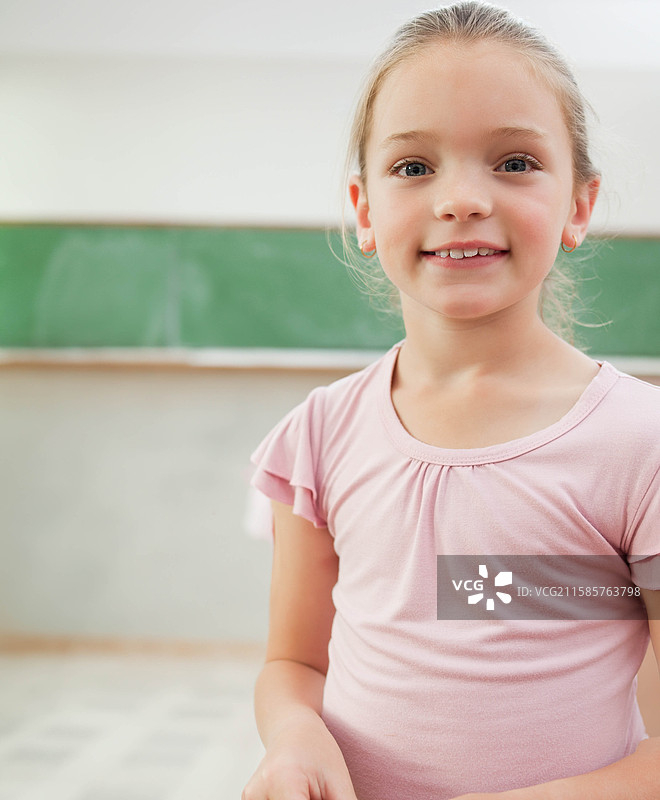微笑的女孩站在瓷砖地板上，面前是绿色黑板，穿着粉色上衣，留有空白空间。图片素材