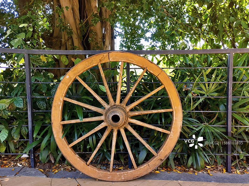 古朴的木制车轮，倚靠在金属围栏上，被绿植与大树环绕，透着岁月沉淀的韵味 。图片素材