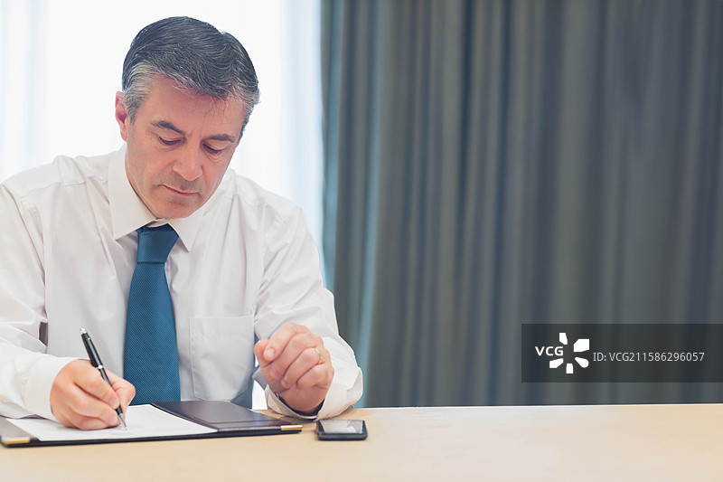 男性穿着白衬衫和蓝色领带，在办公桌上签署文件，手边有智能手机，留有空白空间图片素材