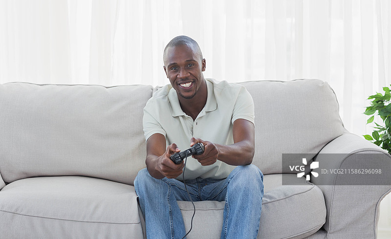 非裔美国男性握着游戏手柄，靠在家里的沙发上，窗帘和植物环绕。图片素材