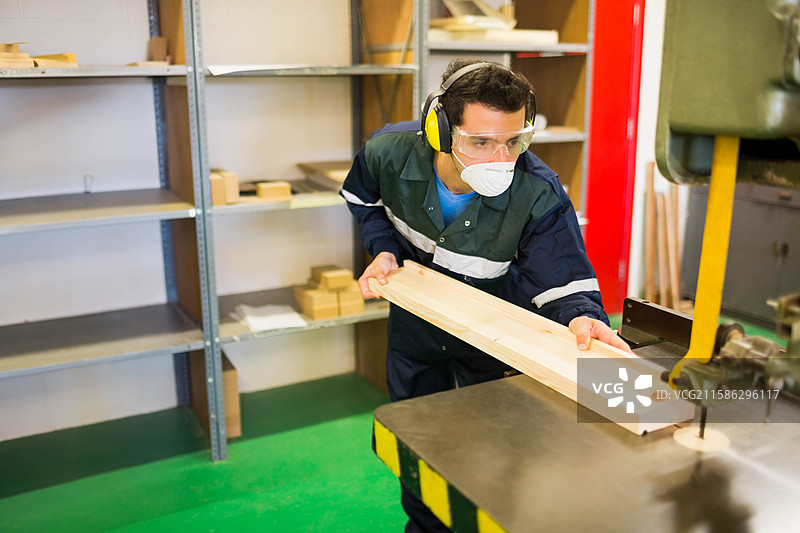穿着工作服和安全装备的男性木工在车间将木板送入带锯。图片素材