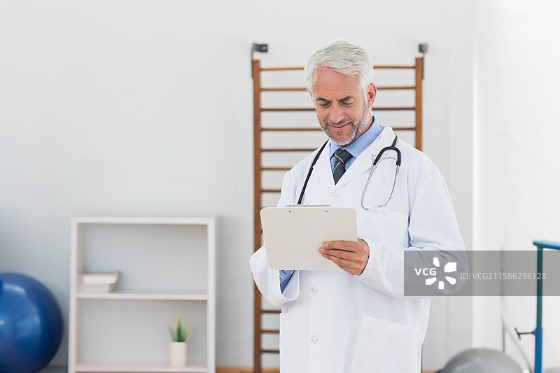 一位年长男性医生站着在康复室阅读夹板上的笔记，佩戴着听诊器，留有空白空间。图片素材