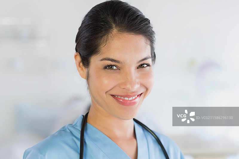 微笑的女性医疗工作者穿着蓝色护士服，站在临床室内，佩戴听诊器。图片素材