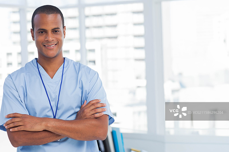 非裔美国男性站在诊所的窗边，穿着浅蓝色的护士服和挂绳证件。图片素材