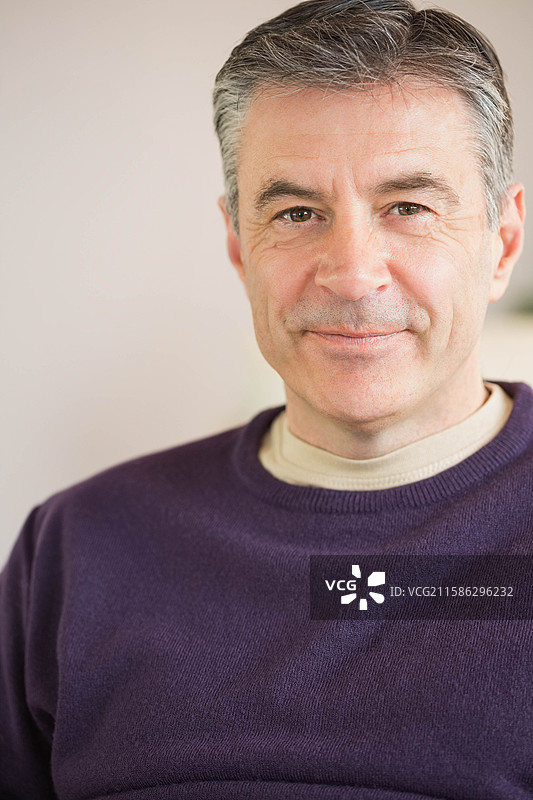 中年男子在光线明亮的房间里摆姿势，穿着紫色毛衣和米色衬衫，背景有模糊的植物。图片素材