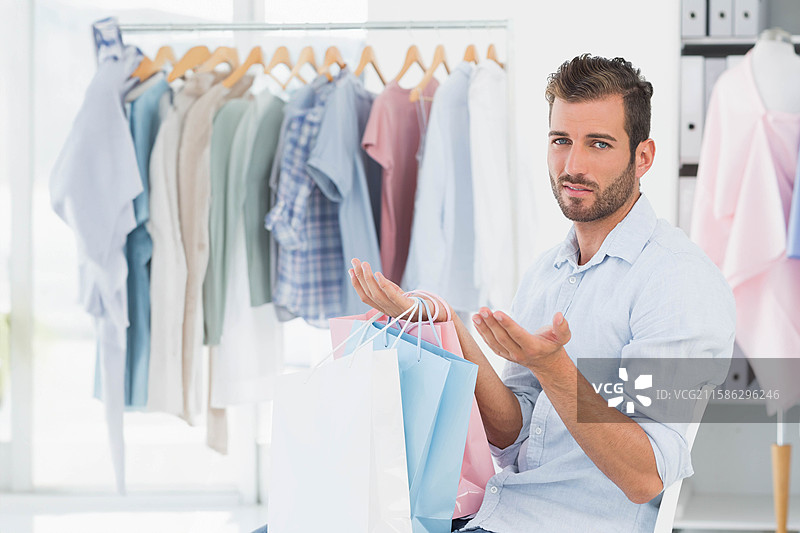 男子坐在服装店里，手里拿着淡色购物袋，正在靠近衣架和模特做手势。图片素材