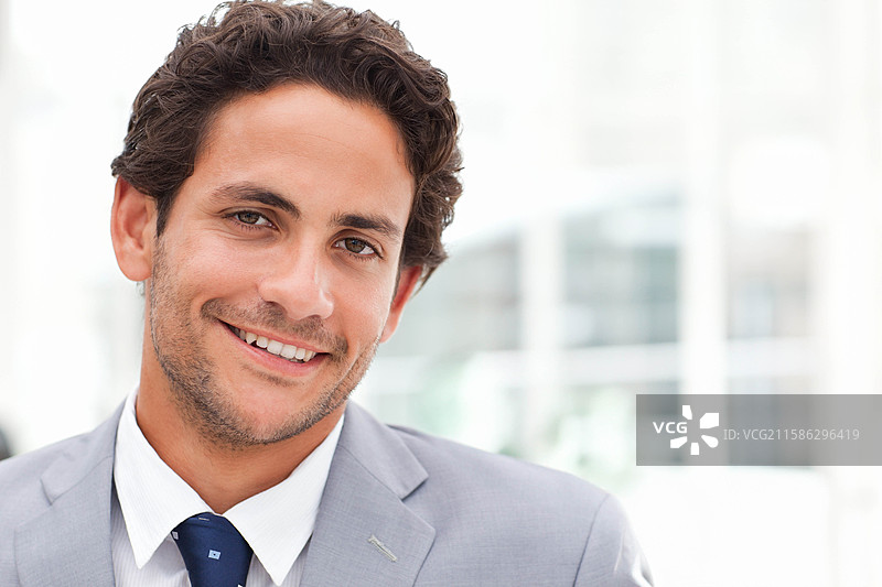 微笑的男性半身像，穿着浅灰色西装和深蓝色领带，背景为模糊的办公室。图片素材