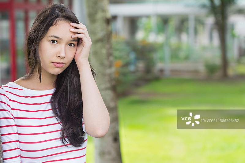 二十多岁的女性站在树干旁，轻触头发，身处红色栏杆的庭院中，留有空白空间。图片素材