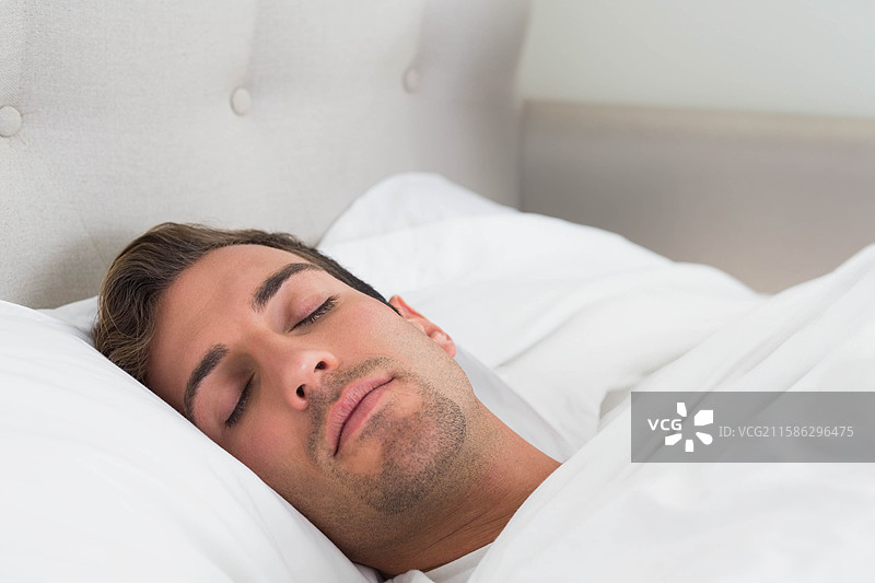 男人躺在白色床单下，头枕在白色枕头上，背靠米色床头板，身处卧室中。图片素材