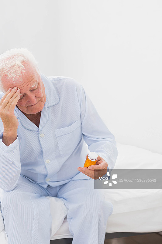 穿着睡衣的老年男子坐在卧室床边，手握药瓶，揉着太阳穴，留有空白空间。图片素材