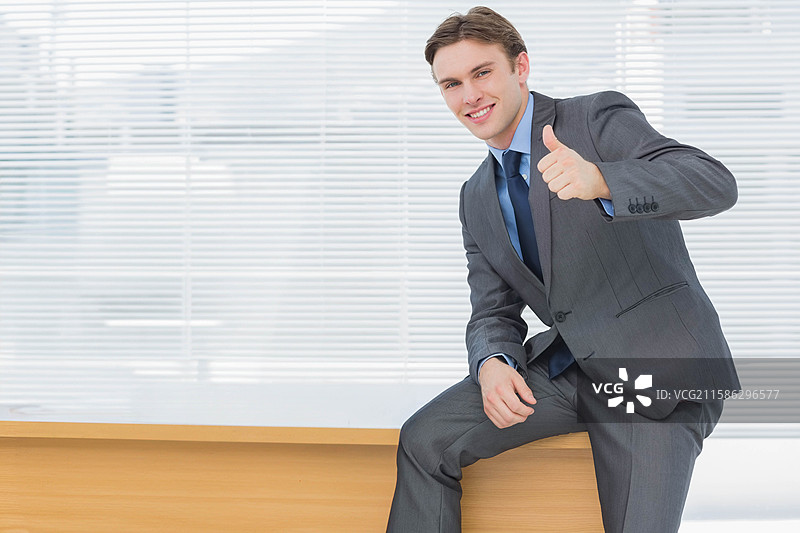 穿着西装和领带的男子坐在办公室的长椅上，靠在水平百叶窗旁，竖起大拇指。图片素材