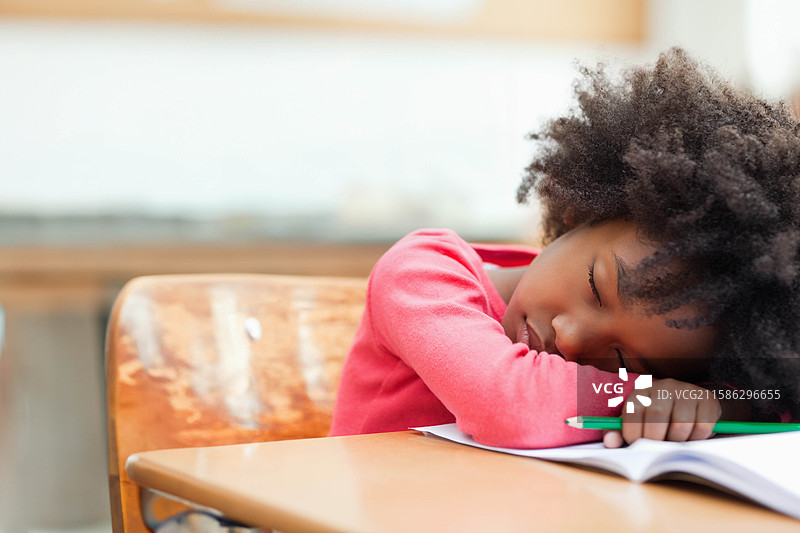非裔美国女孩将头靠在桌子和椅子上，手握绿色铅笔，专注于课本。图片素材
