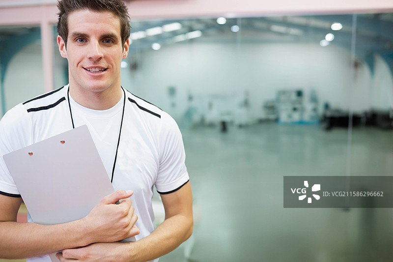 男教练手持夹板，佩戴哨子，站在健身房的玻璃隔断旁，留有空白空间。图片素材