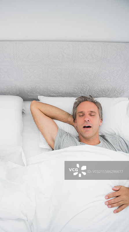 男人在卧室的床上躺着，休息在白色的被子下，枕头和绗缝床头板陪伴着他。图片素材