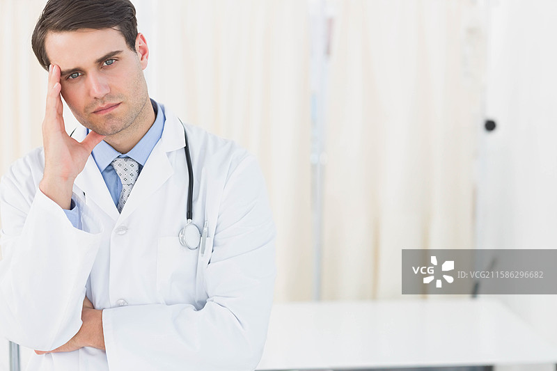 男医生穿着白大褂，沉思地站在检查室里，旁边有听诊器，留有空白空间。图片素材