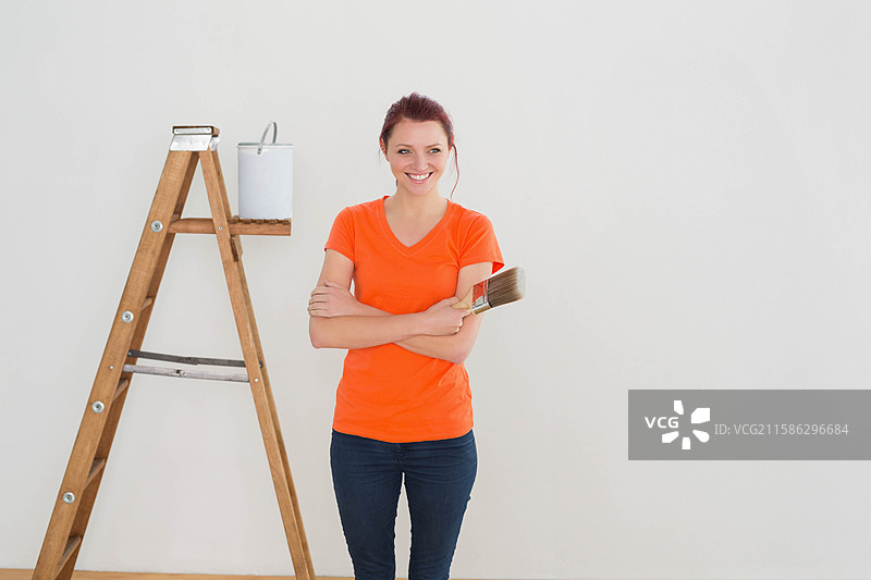 女人站在空房间里，手持画笔和油漆桶，旁边有一把梯子，面前是一面空白的墙。图片素材