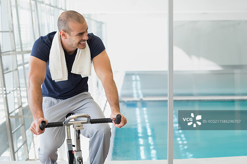 在健身房骑动感单车的男子，旁边有隔断，俯瞰游泳池，身上披着白色毛巾，留有空白空间。图片素材