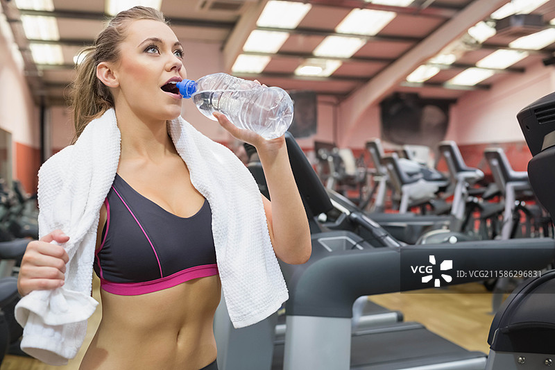 女性在明亮的健身房中暂停跑步机锻炼，喝着透明塑料瓶里的水图片素材
