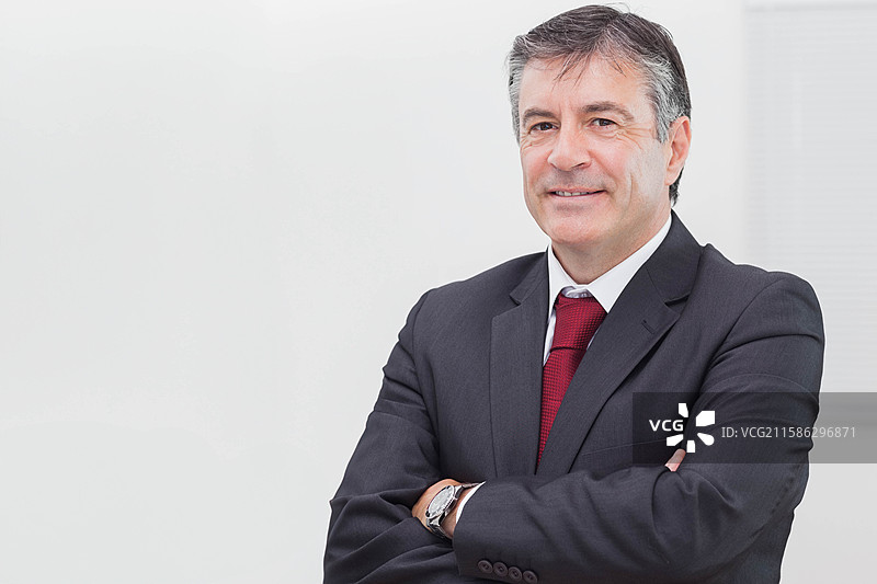 中年男性高管在办公室交叉双臂，身穿西装和红色领带，手腕上戴着手表，留有空白空间图片素材