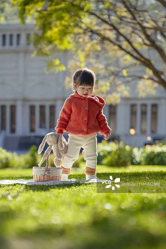 日光下博物馆树前草坪上手拿布偶玩乐的可爱小女孩图片素材