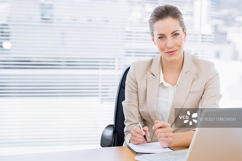 穿着西装的女商人在办公桌上用笔写笔记本，旁边有笔记本电脑，留有空白空间。图片素材