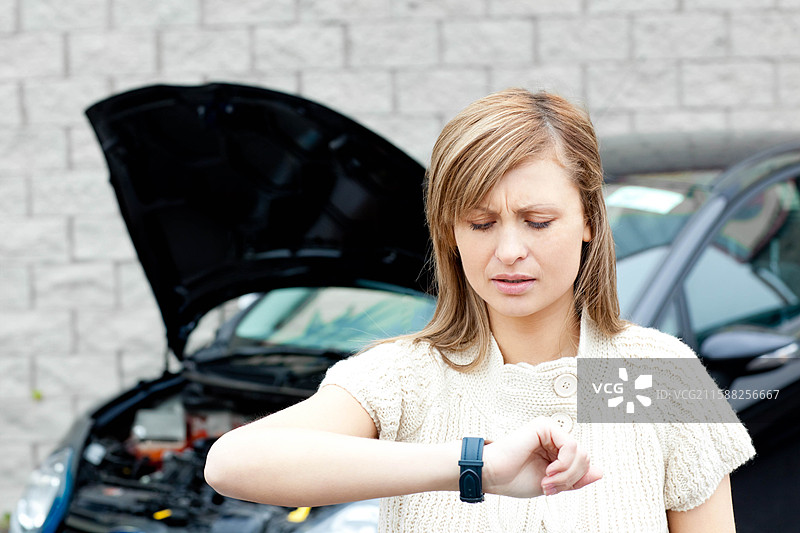 女人站在石墙旁边的故障汽车旁，查看智能手表，汽车引擎盖打开。图片素材
