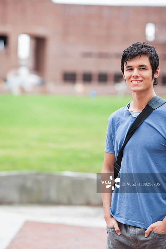 男学生背着书包站在人行道上，旁边是绿草坪和砖砌建筑，留有空白空间。图片素材