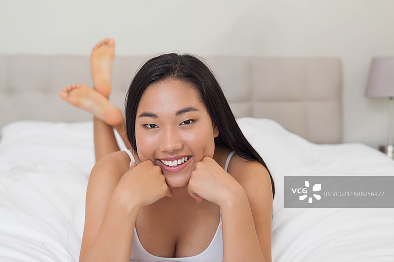 亚洲女性躺在卧室的床上，用双手托着下巴，白色的被子和薰衣草色的台灯。图片素材