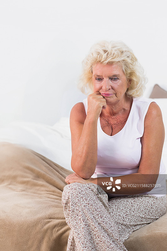 一位年长女性坐在柔和灯光的卧室床边，凝视着米色的毯子，留有空白空间。图片素材